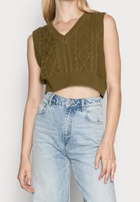 Sleeveless olive green cable knit cropped sweater vest paired with high-waisted light blue acid-wash jeans on a standing person.
