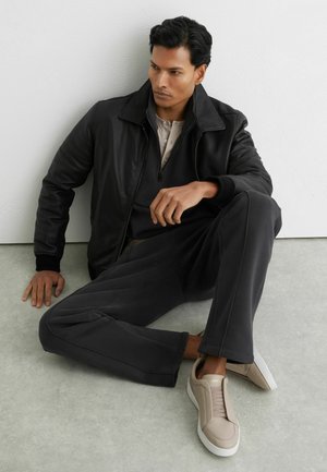 Mann in schwarzer Lederjacke, dunkler Hose, beigen Sneakern, sitzt auf dem Boden vor weißer Wand und schaut mit ernster Miene zur Seite.