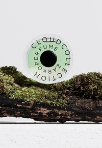 Grønn glass sirkel-formet parfymeflaske med en svart hette, merket "CLOUD COLLECTION", plassert på mosekledd tre.