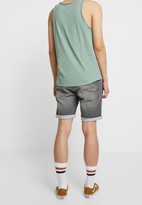 Camiseta sin mangas para hombre en verde claro, combinada con pantalones cortos de mezclilla gris, con dobladillos enrollados, calcetas blancas con rayas rojas y negras, y zapatillas marrones.