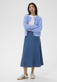Femme debout, les bras croisés, portant un cardigan en tricot bleu clair, un t-shirt blanc, une jupe midi en denim et des baskets blanches avec des rayures noires.