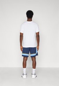 T-shirt branca de mangas curtas e calções azul-marinho com riscas azuis e amarelas. Meias brancas e sapatilhas desportivas completam o conjunto.