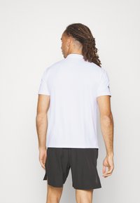 Biela polokošeľa s krátkym rukávom, mäkký materiál, štandardný golier, minimalisticý dizajn. Nosená s čiernymi šortkami, ktoré vykazujú uvoľnený strih a bočné rozparky.