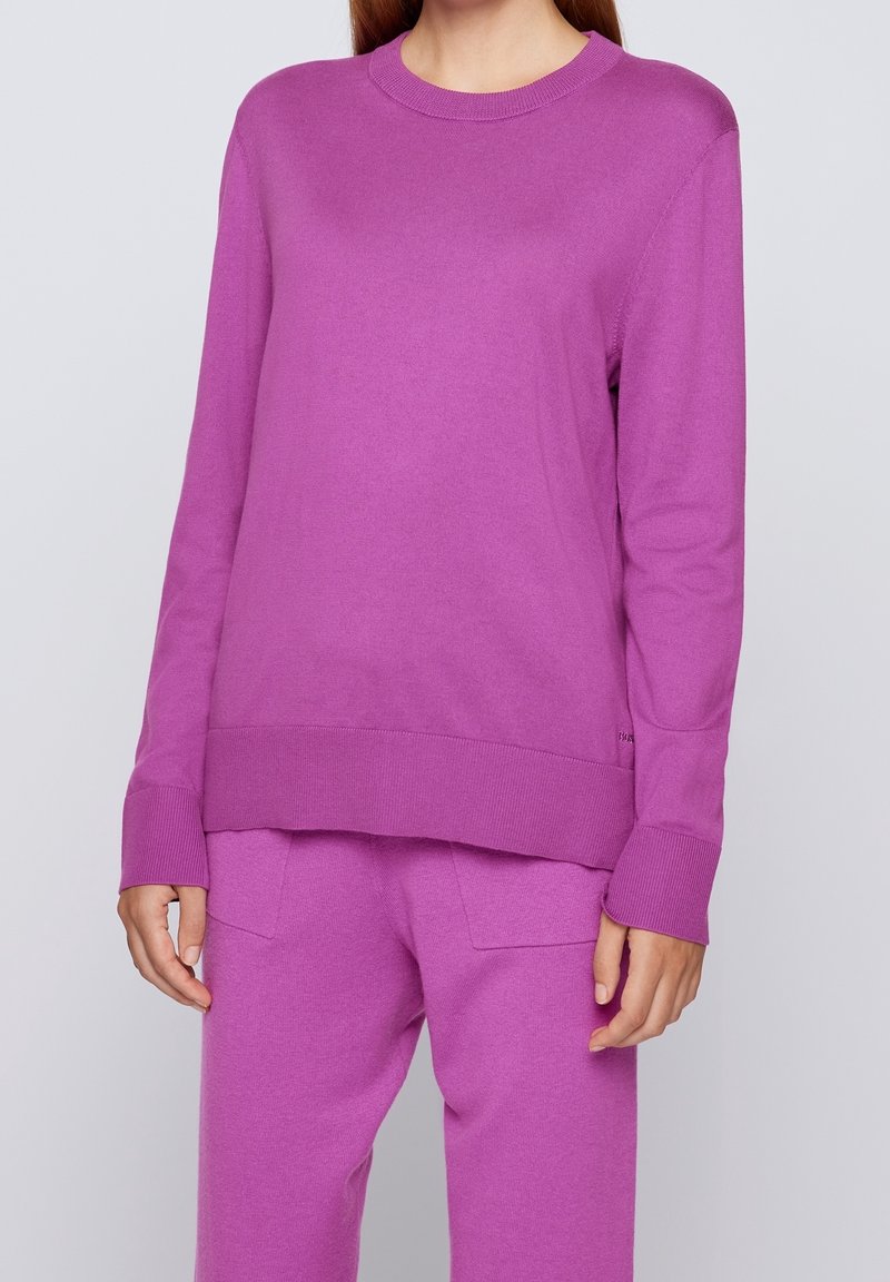 Kvinna i en matchande ljuslila stickad tröja och byxset, stående mot en enkel grå bakgrund.