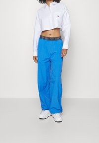 Avskuren vit skjorta med bröstficka; blå löst sittande byxor med svart elastiskt midjeband märkt "TOMMY JEANS"; vita skor.