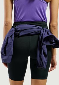 Zwarte fietsbroek gecombineerd met een paarse tanktop, voorzien van een geknoopte, losse paarse jas die om de taille is geknoopt. Glad stoftextuur zichtbaar.
