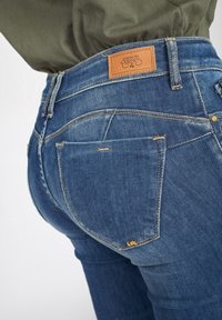 Jeans de mezclilla azul con un ajuste slim, que cuentan con una etiqueta de cuero marrón en la cintura y un desvanecimiento sutil en la tela.