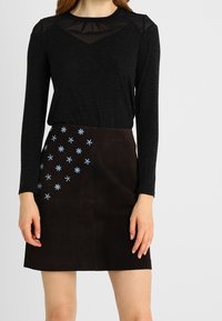 Černý top s dlouhým rukávem a průhledným výstřihem, spárovaný s hnědou sukní z nubuku, která má na jedné straně modrou květinovou výšivku.