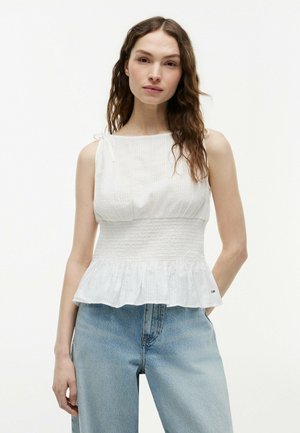 Vrouw met lang golvend bruin haar draagt een witte mouwloze gerimpelde top en lichtblauwe high waist jeans tegen een effen witte achtergrond.