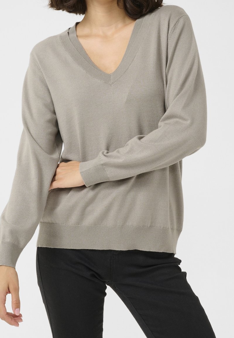 Femme portant un pull léger taupe à col en V et un pantalon noir, debout devant un fond neutre.