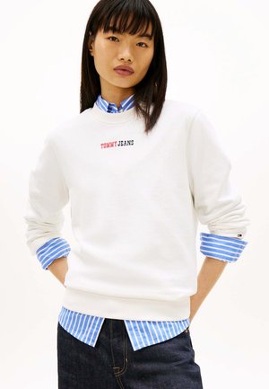 Giovane donna che indossa una felpa bianca Tommy Jeans sopra una camicia a righe blu e bianca, abbinata a jeans scuri, con le mani in tasca.