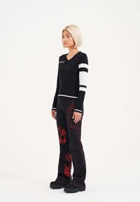 Black sweater with white striped sleeves, V-neck design. Black pants featuring red graphic patterns and flared legs. Chunky black footwear.