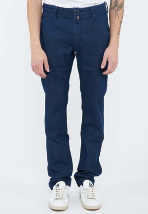 Man wearing dark blue straight-fit pants with white sneakers, standing against a plain background.