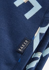 Marinblått tyg med geometriska mönster i ljusblått och krämfärgat. Har en svart etikett med "BAKER" och "stylad rätt" i vit text.