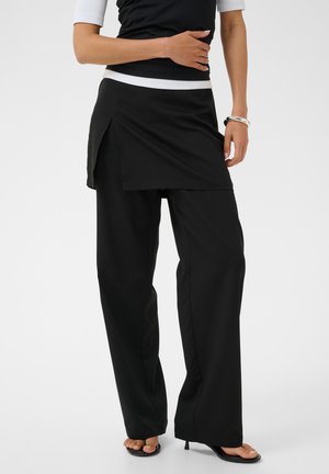 Personne portant un pantalon noir à jambes larges avec une jupe superposée assortie, un haut blanc à manches courtes et des sandales à talons noires.