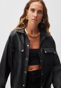 Giacca in ecopelle nera con due tasche sul petto, dettagli dorati e una vestibilità rilassata. Abbinata a un top nero per un look stratificato.