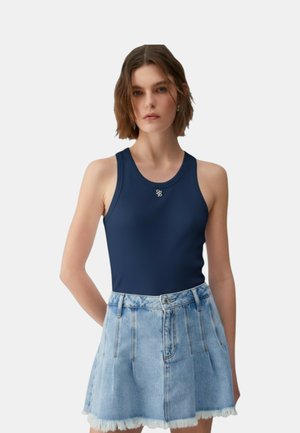 Donna che indossa una canottiera a coste blu navy e una gonna in denim azzurro chiaro con orlo sfrangiato, in piedi con le braccia dietro la schiena.