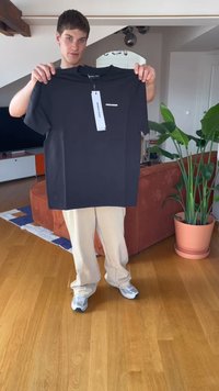 Melns T-kreklis, kas izgatavots no kokvilnas, ar apaļu kakla izgriezumu un īsām piedurknēm, ar baltu logotipu uz krūtīm un pievienotu taisnstūrveida cenu birku.