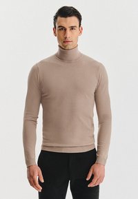 Beżowy sweter z golfem wykonany z miękkiego dzianinowego materiału. Dopasowany fason z prążkowanymi mankietami i dołem, noszony z ciemnymi spodniami. Gładka faktura.