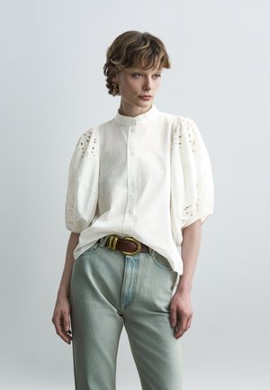 Nő fehér, gombos blúzban, melynek fodros, kivágott ujja van, világoskék farmert visel, és barna övet arany csattal.