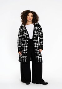 Schwarz-weißer gemusterter langer Cardigan mit gebundenem Taillenbund, langen Ärmeln und lockerer Passform, getragen über einem schlichten weißen Oberteil und weiten schwarzen Hosen.