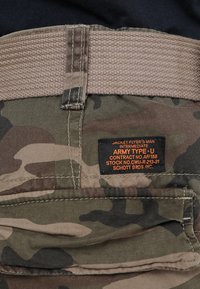 Camo-broek met een beige geweven riem. Voorzien van een zwart label met oranje tekst die merk- en productinformatie vermeldt. Gelaagd stof.