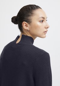 Dunkelblaues Strickpullover mit hohem Kragen und Schlitzdetail auf der Rückseite, ausgestattet mit gerippter Struktur und körpernahem Schnitt.