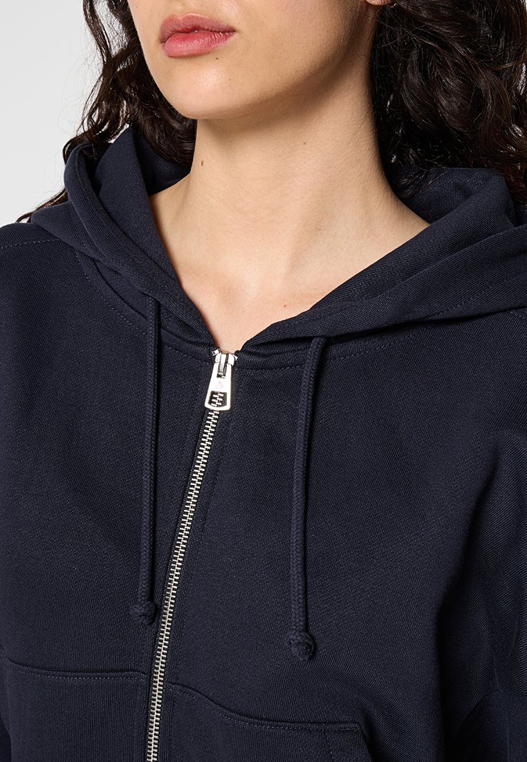 Granatowa bluza z kapturem na zamek wykona z teksturowanej tkaniny; posiada kieszeń z przodu, sznurek i srebrny metalowy zamek.
