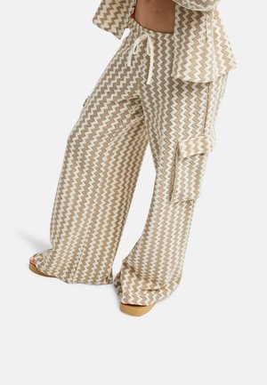 Spodnie o szerokich nogawkach z beżowo-białym wzorem w zygzak, boczną kieszenią typu cargo, ściągaczem w pasie, noszone z pasującą wzorzystą kurtką i beżowymi sandałami.