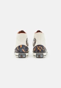 Białe kanwasowe botki za kostkę z brązowym obcasem w leopardzi wzór, posiadające czarną naszywkę z logo Converse oraz klasyczną gumową podeszwę w paski.