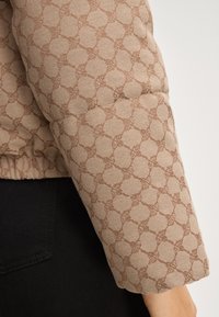 Beżowa pikowana kurtka z teksturowanym, powtarzającym się wzorem przeplatających się kształtów. Tkanina wydaje się być miękka z lekko błyszczącym wykończeniem.