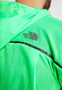 Veste à capuche vert vif avec logo "The North Face" et bande noire sur le haut du dos portée par une personne.