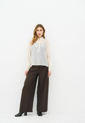 Femme aux longs cheveux portant un chemisier blanc transparent avec un nœud et un pantalon large marron foncé fluide, ainsi que des bottines noires.
