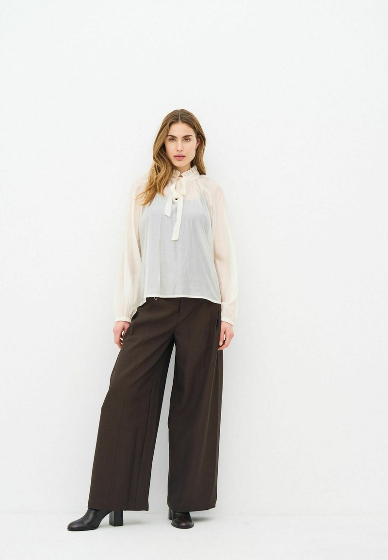 Femme aux longs cheveux portant un chemisier blanc transparent avec un nœud et un pantalon large marron foncé fluide, ainsi que des bottines noires.