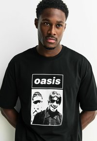 Tricou negru din bumbac cu o grafică albă ce înfățișează două figuri purtând ochelari de soare. Textul "oasis" deasupra imaginii. Croi obișnuit.