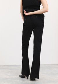 Pantaloni neri su misura con gamba svasata, caratterizzati da una texture liscia e due tasche posteriori con chiusura a bottone. Indossati con scarpe nere con tacco.