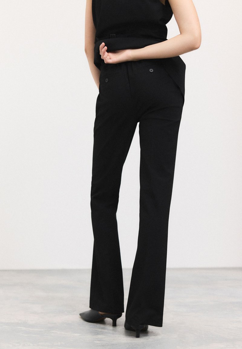 Pantaloni neri su misura con gamba svasata, caratterizzati da una texture liscia e due tasche posteriori con chiusura a bottone. Indossati con scarpe nere con tacco.