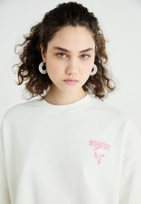 Witte sweatshirt met "HOWDY" in roze tekst en grafiek, ronde strassoorbellen. Zacht stof, ontspannen pasvorm en rondenek ontwerp.