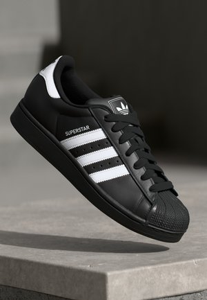 Baskets Adidas Superstar noires avec des bandes blanches et embout coquille, montrées en plein air au-dessus d'une surface en béton.