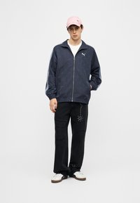 Νέος άνδρας φοράει ροζ καπέλο, μπλε σκούρο μπουφάν με φερμουάρ και λογότυπο, λευκά αθλητικά παπούτσια και μαύρο παντελόνι με φθαρμένες λεπτομέρειες και σχέδιο με τελείες, στέκεται μπροστά σε λευκό φόντο.