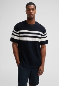 Marineblauer Strickpullover mit weißen und grauen horizontalen Streifen, rundem Halsausschnitt und kurzen Ärmeln. Kombiniert mit dunklen Shorts.