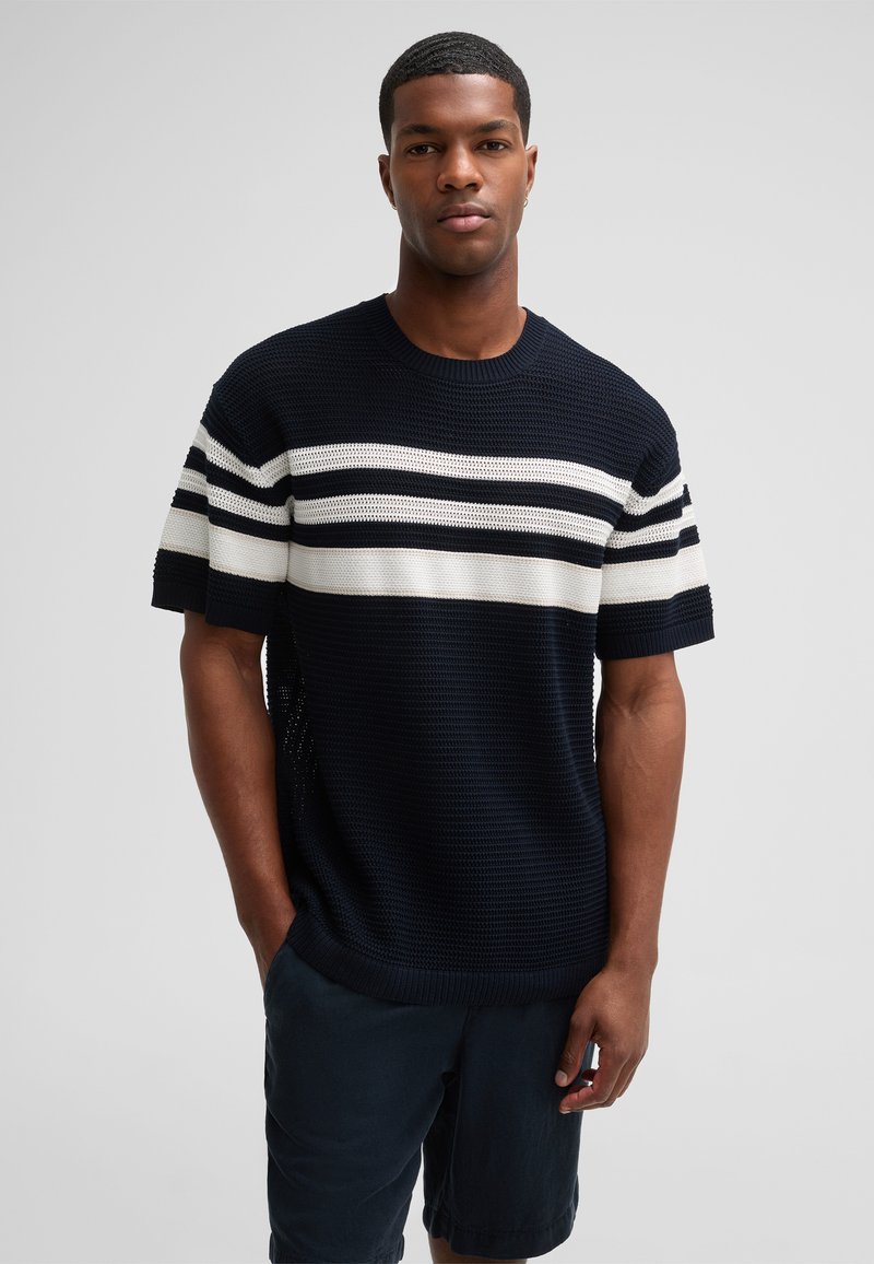 Marineblauer Strickpullover mit weißen und grauen horizontalen Streifen, rundem Halsausschnitt und kurzen Ärmeln. Kombiniert mit dunklen Shorts.