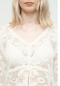 Γυναίκα με ξανθά μαλλιά μήκους μέχρι τους ώμους, που φορεί ένα λευκό πλεκτό ζακέτα με λουλουδάτα σχέδια, κουμπιά μπροστά και κορδόνι δεμένο στη μέση.