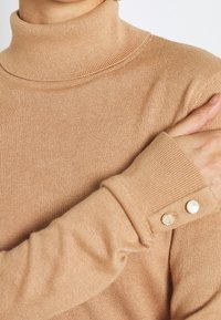 Hnědý rolák ze měkké pleteniny, který má ribbovanou strukturu a manžety s knoflíky zdobené kovovými akcenty.
