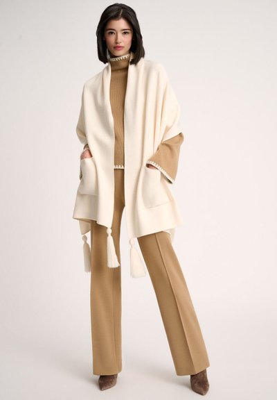 Cape de couleur crème avec des franges, associée à un pull en maille brun et un pantalon large de couleur tan. Tissu doux et silhouette structurée.