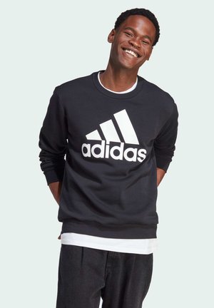 Sudadera negra con el logo blanco de Adidas, cuello redondo y mangas largas. Lleva puesta sobre una camisa blanca, con tejido texturizado y un ajuste relajado.