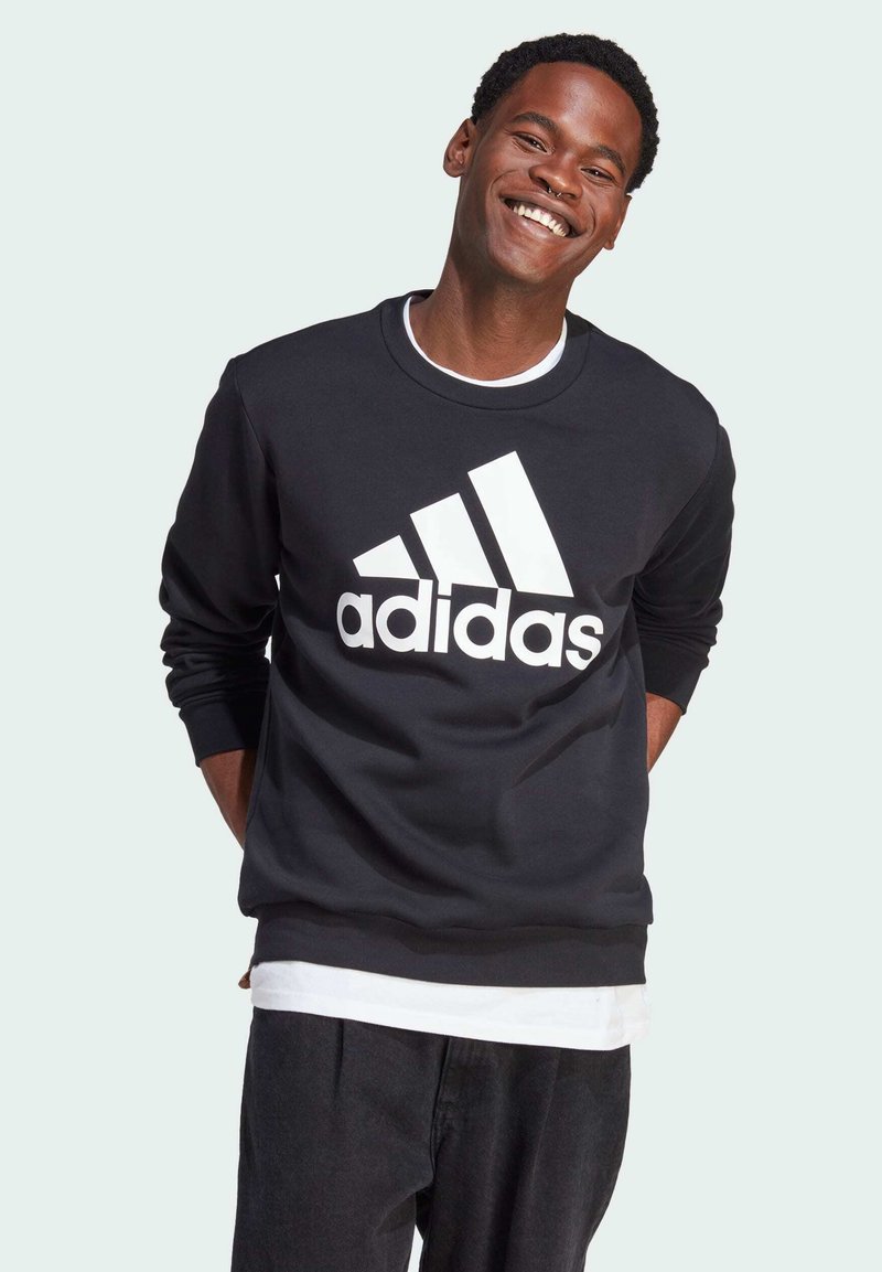 Sudadera negra con el logo blanco de Adidas, cuello redondo y mangas largas. Lleva puesta sobre una camisa blanca, con tejido texturizado y un ajuste relajado.