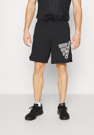 Homme portant un short Adidas noir avec un grand logo blanc sur la cuisse droite, baskets noires, chaussettes blanches, debout sur fond blanc.