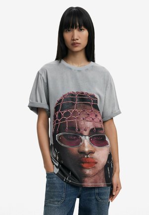 T-shirt gris à manches courtes avec un motif de visage imprimé, des accents roses, des lunettes brillantes et un accessoire de tête texturé.