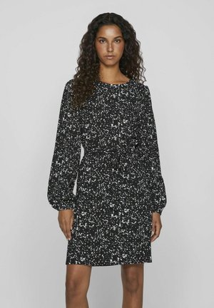 Robe noire avec un motif blanc éparpillé, encolure ronde, manches longues ballons et un détail de taille nouée. Texture du tissu lisse.
